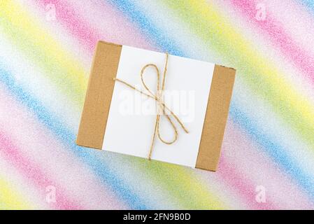 Ein kleiner Karton aus Wellpappe, umwickelt mit einem weißen Blatt und einer grauen Schnur in einer Schleife, isoliert auf einem bunten Hintergrund, Draufsicht. Stockfoto