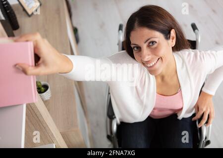 Frau im Rollstuhl, die aus dem hohen Regal nach Büchern greift Stockfoto