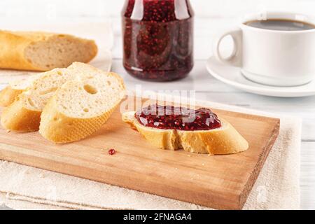 Leckeres Sandwich mit süßer Himbeermarmelade und Baguette-Scheiben auf einem Holzschneidebrett gegen Marmeladenglas und Tasse Kaffee über dem Holztisch. Frühstück. Stockfoto