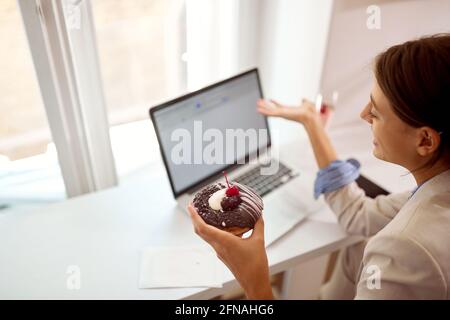 Eine junge Geschäftsfrau sieht sich den Inhalt eines Laptops an Eine angenehme Atmosphäre in einer Konditorei, während ein Köstliche Donut in ihrer Hand, dass sie Stockfoto