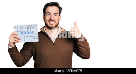 Junger hispanischer Mann hält den Reisekalender glücklich und positiv, Daumen hoch und tut ausgezeichnet und Zustimmung Zeichen Stockfoto