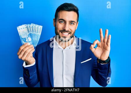 Der junge hispanische Geschäftsmann trägt einen Business-Anzug mit chilenischen Pesos-Banknoten und tut ein gutes Zeichen mit den Fingern Stockfoto