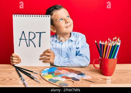 Entzückender kaukasischer Kindermaler, der im Kunststudio sitzt und ein lächelndes Künstlerwort hält und zur Seite schaut und dabei denkt. Stockfoto