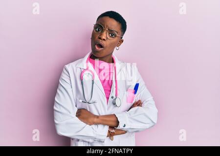 Junge afroamerikanische Frau in Arztuniform und Stethoskop im Schockgesicht, skeptisch und sarkastisch aussehend, überrascht mit offenem Mund Stockfoto