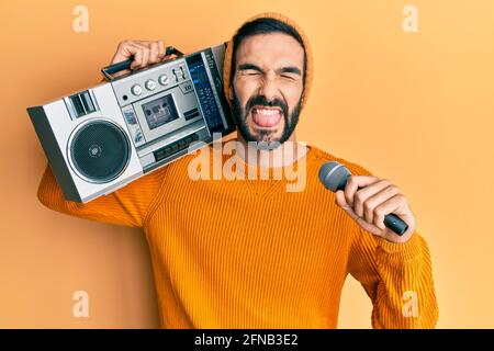 Junger hispanischer Mann hält eine Boombox und hört Musik, die mit einem Mikrofon singt, das die Zunge aus dem Mund ragt, glücklich mit einem lustigen Ausdruck. Stockfoto