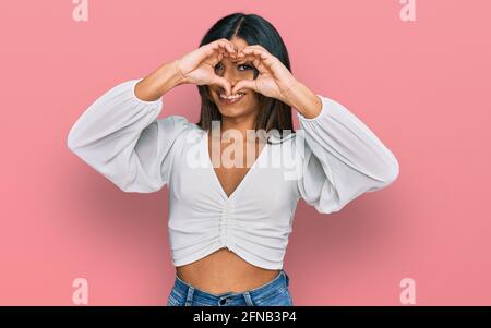 Junge transsexuelle lateinische Transgender-Frau in lässiger Kleidung mit Herz Form mit Hand und Finger lächelnd Blick durch Zeichen Stockfoto