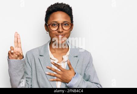 Junge afroamerikanische Frau in Geschäftskleidung lächelnd, mit der Hand auf der Brust und den Fingern nach oben verfluchend, einen Treueversprechen-Eid abmachend Stockfoto