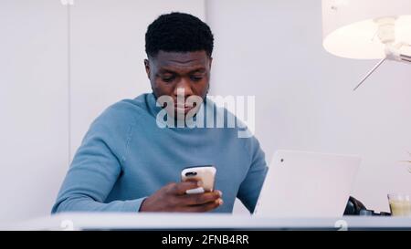 afroamerikanischer schwarzer Mann mit ernstem Gesichtsausdruck, der das Telefon vor dem Laptop benutzt. Hochwertige Fotos Stockfoto