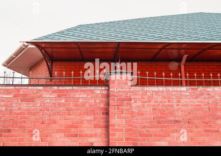 Die Mauer des Zauns besteht aus rotem Backstein, das Dach des Hauses ist von hinten sichtbar. Stockfoto