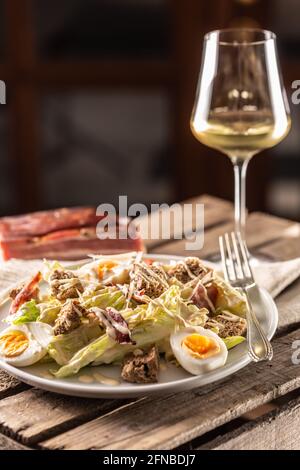 Caesar-Salat mit gekochten Eiern, Speck und Croutons, serviert auf einem Teller neben einem Glas Weißwein. Stockfoto