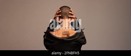 Rückansicht einer Genderqueerin, die ihre Ohren auf grauem Hintergrund bedeckt. Frau mit rasierten Kopf und drücken Sie sich auf ihrem Rücken geschrieben. Stockfoto