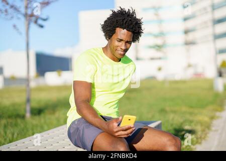 Schwarzer Mann, der sein Smartphone mit einer Übungs-App konsultiert, während er sich von seinem Training ausruhte. Stockfoto