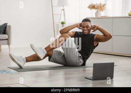 Happy Black Man Macht Elbow-To-Knee Abs Crunches Bei Laptop Indoor Stockfoto