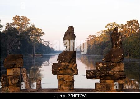 Nahaufnahme einer der Statuen, die den Damm über dem Graben zum Südtor von Angkor Thom säumen. Der Graben ist im frühen Morgenlicht flach ruhig Stockfoto