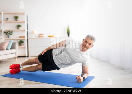 Gesundes Lifestyle-Konzept. Fit reifen Mann steht in der Seitenwand auf Sportmatte zu Hause, Training im Wohnzimmer Stockfoto