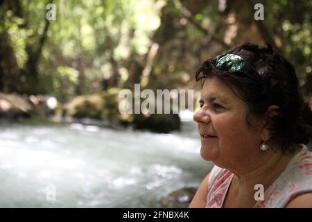 Profil einer lächelnden 62-jährigen älteren Frau im Wald mit dem plätschernden Fluss Banias, einer der drei Quellen des Jordan. Stockfoto