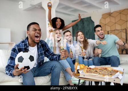 Emotionale multirassische Freunde, die zu Hause Fußball spielen, das Tor schreiend, den Sieg feiern Stockfoto