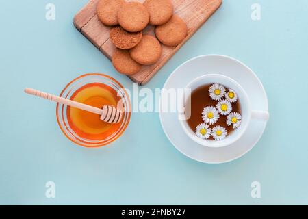 Eine Tasse Tee und Honig in einer Glasschüssel mit Holzdipper und Keksen auf Schneidebrett auf blauem Hintergrund. Gesundes Frühstück. Draufsicht, flach liegend Stockfoto