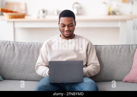 Lächelnder schwarzer Mann, der einen Laptop benutzt und auf dem Sofa sitzt Stockfoto