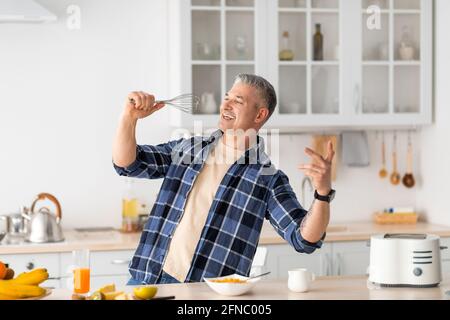 Ein älterer Mann, der beim Kochen des Frühstücks in der modernen Küche in einen Schneebesen getäuscht und gesungen hat. Stockfoto