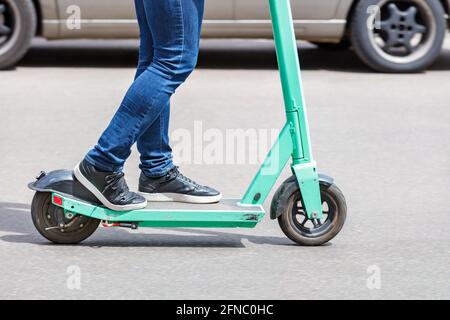 Ein Mann in blauer Jeans fährt an einem sonnigen Tag mit einem vorbeifahrenden Auto im Hintergrund einen gemieteten Elektroroller. Stockfoto