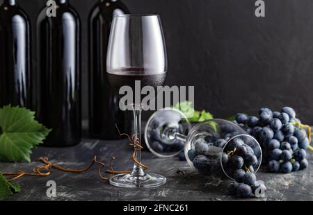 Weinglas mit Rotwein. Rotweinflaschen, Weintrauben mit Blättern und Reben auf dunklem rustikalem Betonhintergrund. Weinkomposition auf schwarzem Stein Stockfoto