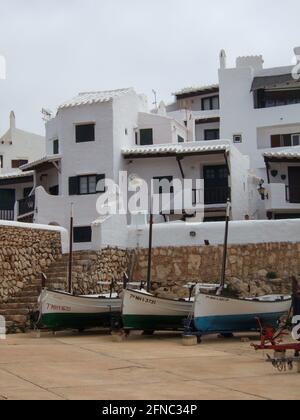 Typisch spanisches weißes Dorf mit einigen weißen Strandhäusern nach kykladischer Architektur auf Menorca. Im Vordergrund sind einige kleine Fischerboote Stockfoto