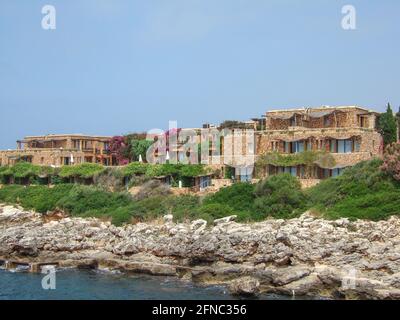 Strandhäuser aus orangefarbenem Sandstein an der felsigen Küste Menorcas. Über der Klippe zum Meer und kleinen bepflanzten Hügeln stehen einige Luxusvillen Stockfoto