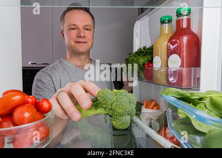 Offener Kühlschrank von innen, Glasregale mit buntem Gemüse, Flaschen mit Bio-Säften. Mann mit einem glücklichen Lächeln, der Brokkoli nahm. Gesunde Ernährung, Stockfoto