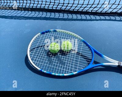 Ein Tennisschläger, der auf zwei gelben Tennisbällen auf einem blauen Platz in der Nähe des Tennisnetzes liegt. Stockfoto