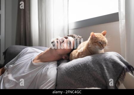 Der junge Mann legt seinen Kopf auf das Sofa daneben Eine braune gestromte Katze und scheint zu träumen Stockfoto