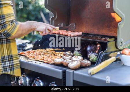 Nahaufnahme eines anonymen Mannes, der Fleisch mit einer Zange auf einem Grill im Freien zubereitet. Hand des jungen Mannes, der einige Spieße, Fleischsteak, Wurst und Gemüse auf einem Gasgrill gegrillt hat, bbq im Garten. Stockfoto