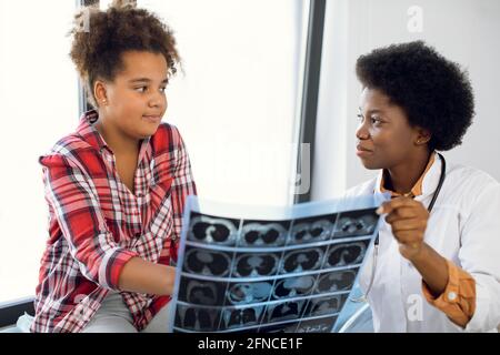 Junge professionelle afroamerikanische Therapeutin, die dem Kind den Röntgenscan erklärt, nettes, interessiertes gemischtes Schulmädchen, das auf der Couch sitzt und den Arzt ansieht. Nahaufnahme der horizontalen Aufnahme Stockfoto