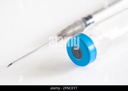 Dünne Spritze hinter einem Corona-Impffläschchen mit blauer Kappe auf weißem Hintergrund. Konzentrieren Sie sich auf die Öffnung an der Nadelspitze, geringe Schärfentiefe Stockfoto