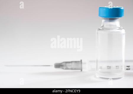 Dünne Spritze hinter einem Corona-Impffläschchen mit blauer Kappe auf weißem Hintergrund. Konzentrieren Sie sich auf die Glasflasche, geringe Schärfentiefe Stockfoto