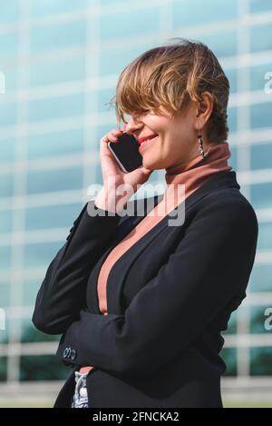 Vertikales Porträt einer attraktiven jungen Frau, die mit dem Telefon spricht. Sie kleidet formelle Kleidung. Bürogebäude im Hintergrund Stockfoto