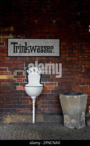 Öffentlicher Brunnen mit weißem Getränk an einer roten Backsteinmauer. Die oben gemalte deutsche Schrift lautet „Wasser trinken“. Stockfoto