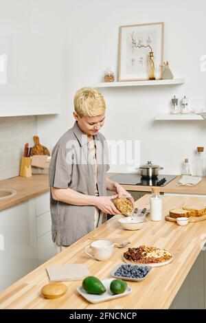 Junge Frau, die am Küchentisch Müsli zum Frühstück zubereitet Stockfoto
