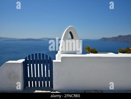 Traditionelle Hausfassade mit einer weiß getünchten Wand, einer blauen Holzzauntür und einem kykladischen Kamin auf Santorini, Griechenland. Stockfoto