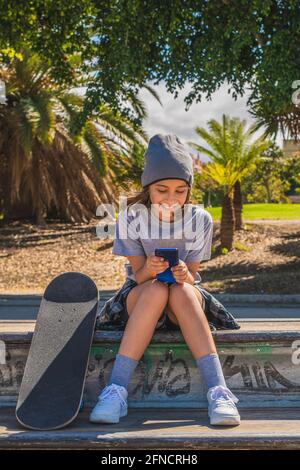 Das Mädchen aus der Vorpubertät, kaukasisch, sitzt auf einem Hindernis in einem Skatepark, mit den Füßen auf ihrem Brett, während sie mit ihrem Mo chattet oder im Internet surft Stockfoto
