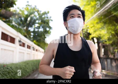 Junger asiatischer Mann, der mit einer Gesichtsmaske in der Stadt läuft. Laufmännchen trainieren und joggen auf der Straße Stockfoto