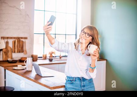 Glückliche reife Frau, die ihren Morgenkaffee trinkt und einen Videoanruf hat, während sie zu Hause in ihrer Küche sitzt. Stockfoto