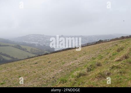 Blick über grasbewachsene Felder an einem grauen Tag: Sanfte Hügel mit Zaun oder Hecke am Horizont entlang und das Meer nur in der nebligen Ferne sichtbar Stockfoto