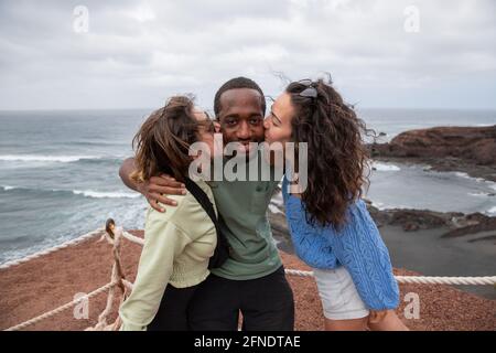 Ein afrikanischer Kerl erhält einen Kuss von zwei kaukasischen Mädchen während eines Urlaubs, eine glückliche multirassische Gruppe macht Spaß Stockfoto