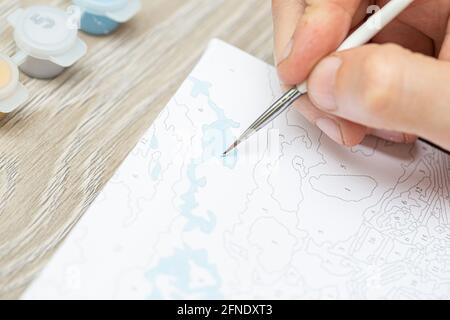 Eine mans Hand hält einen Pinsel und zeichnet ein Bild durch Zahlen. Zeichnen auf Leinwand. Nummerierte Farbdosen. Kreatives Hobby. Malen für Anfänger.Nahaufnahme, sele Stockfoto