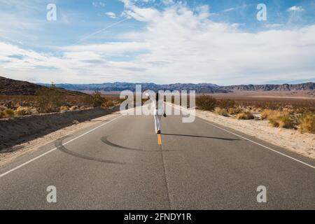 Eine junge Frau, in Weiß gekleidet, die eine trostlose Wüste hinunterläuft Straße in der Mojave-Wüste Stockfoto