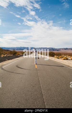 Eine junge Frau, in Weiß gekleidet, die eine trostlose Wüste hinunterläuft Straße in der Mojave-Wüste Stockfoto