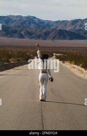 Eine junge Frau, in Weiß gekleidet, die eine trostlose Wüste hinunterläuft Straße in der Mojave-Wüste Stockfoto