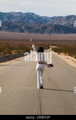 Eine junge Frau, in Weiß gekleidet, die eine trostlose Wüste hinunterläuft Straße in der Mojave-Wüste Stockfoto