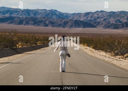 Eine junge Frau, in Weiß gekleidet, die eine trostlose Wüste hinunterläuft Straße in der Mojave-Wüste Stockfoto